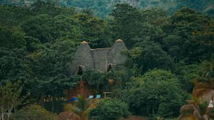 Simple thatched-roof camp buildings surrounded by lush greenery and hills